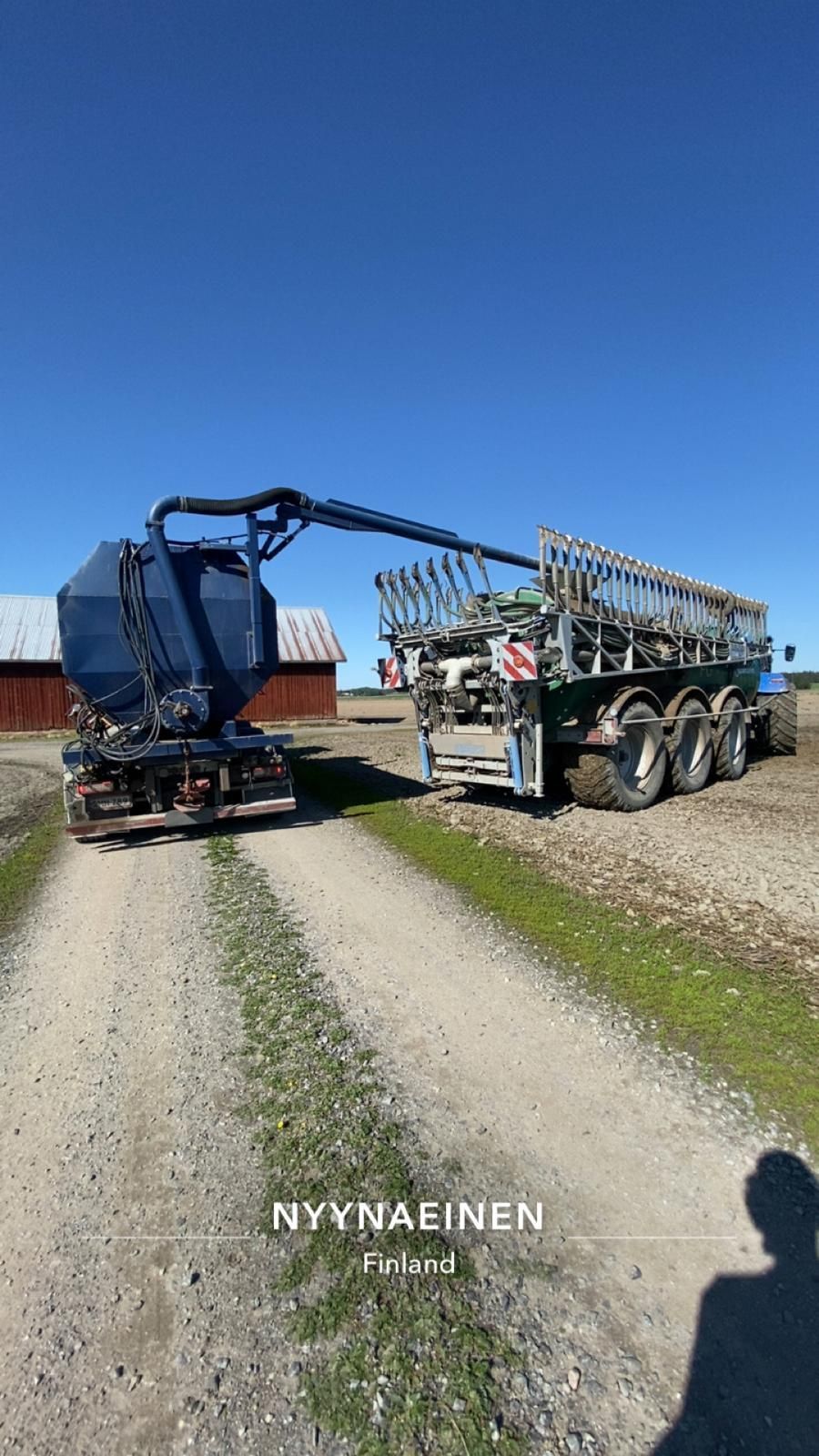 Sininen kuorma-auto, jossa on pitkä varsi, purkaa kuormaa suurempaan vihreään perävaunuun hiekkatiellä aurinkoisena päivänä.