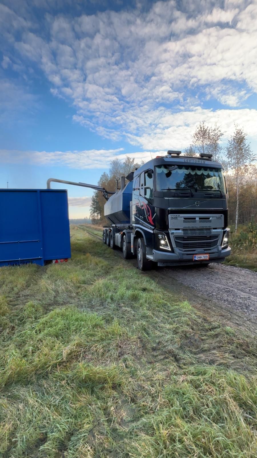 Tumma kuorma-auto, jonka vieressä on säiliö, sinisen rakenteen vieressä, pilvinen taivas yläpuolella.