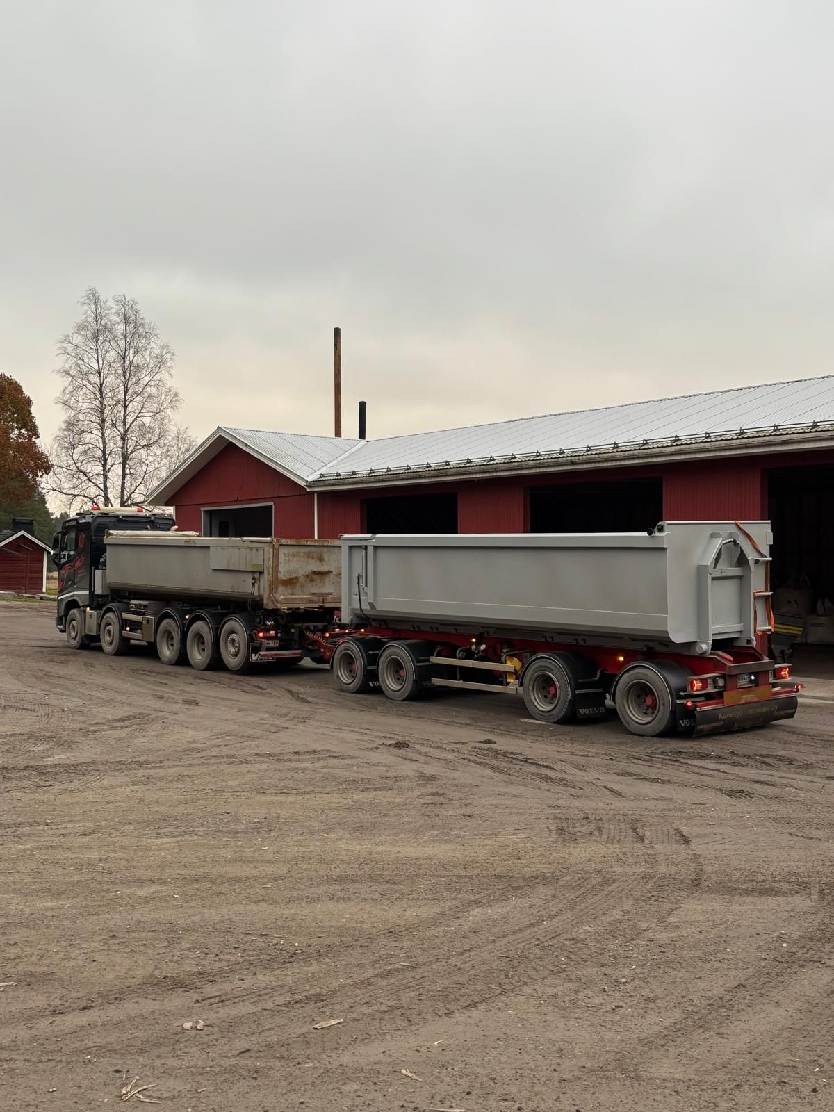 Kahta kippiperävaunua vetävä rekka-auto pysäköitynä punaisen rakennuksen eteen sorakentälle. Pilvinen taivas.