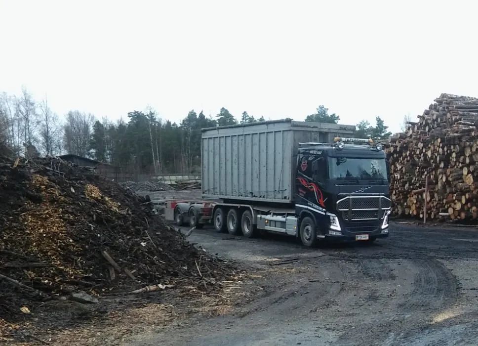 Musta rekka vetää suurta konttia puunjalostusalueella; puu- ja tukkikasat näkyvät.