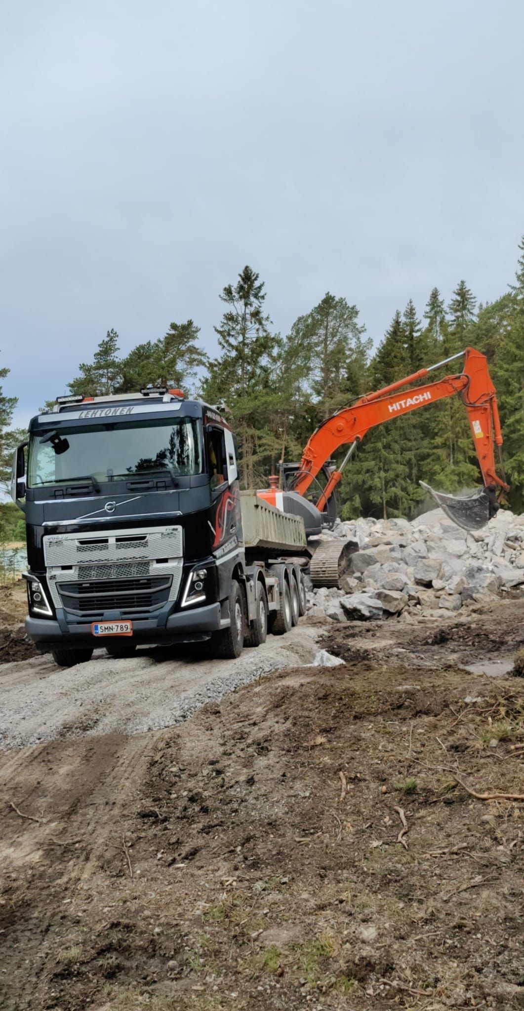 Musta kuorma-auto kaivinkoneella lastaa kiviä hiekkatiellä metsän vieressä.