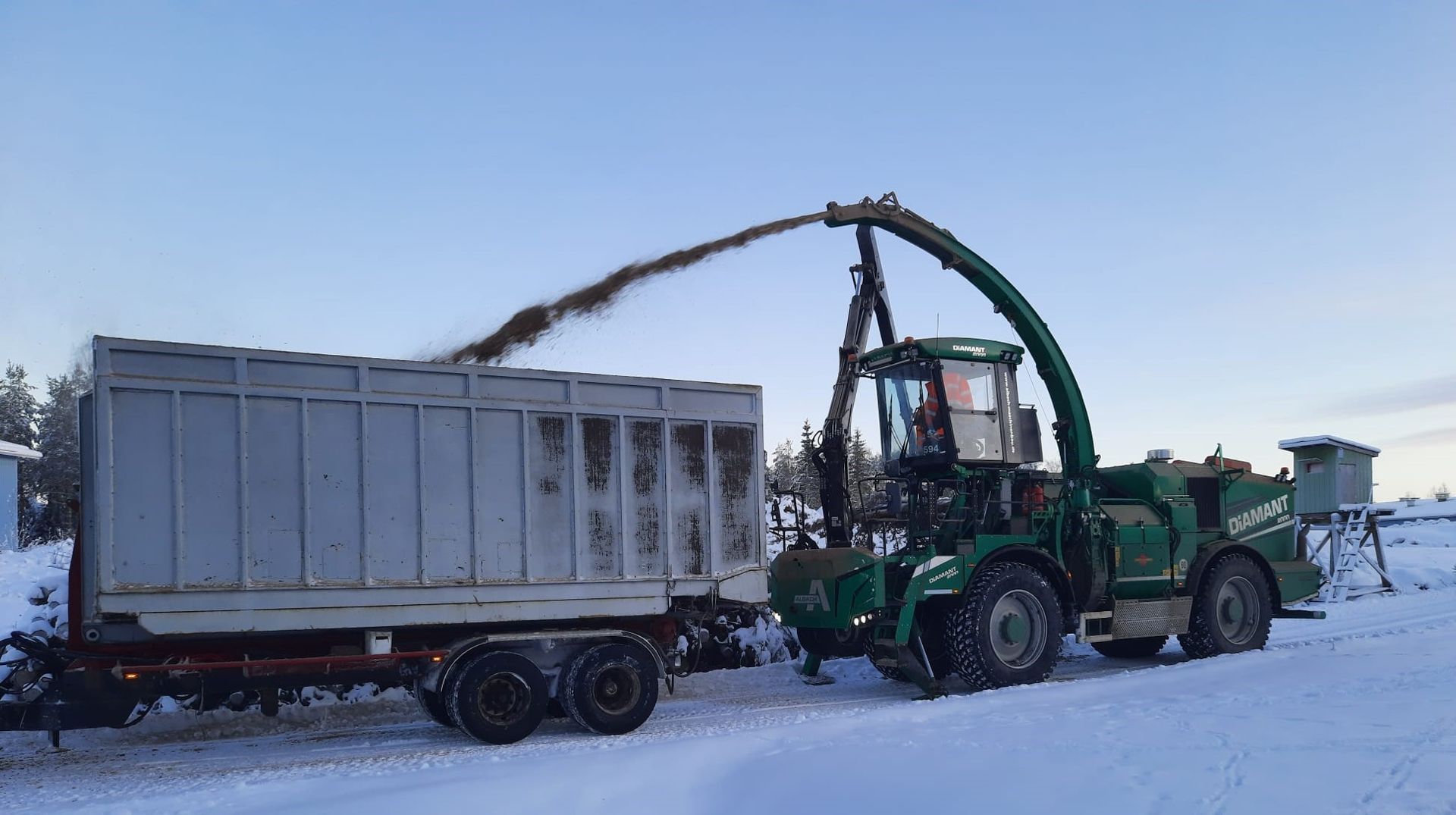 Vihreä hakkuri lastaa perävaunua puuhakkeella lumisella maisemalla.