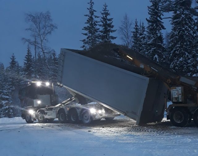 Kuorma-auto kippaa lastia lumisessa metsässä hämärässä.