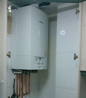 Una caldera de gas blanca montada en la pared, encerrada dentro de un mueble de cocina, que muestra tuberías de cobre expuestas debajo.