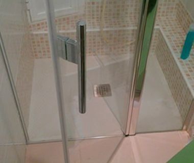 Una ducha a ras de suelo con puerta de cristal, manija de metal y desagüe cuadrado, sobre una pared de azulejos cuadrados pequeños de colores claros.