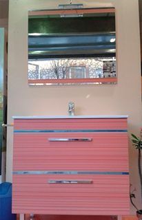 Un tocador de baño de dos cajones de color coral con encimera blanca, herrajes cromados y un espejo rectangular a juego.