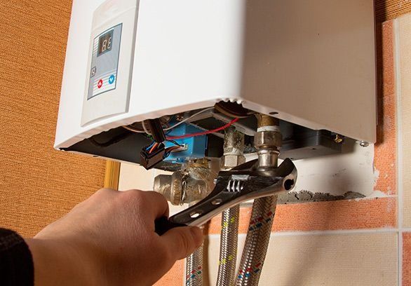 Una mano utiliza una llave ajustable para apretar una conexión metálica en un calentador de agua montado en la pared.