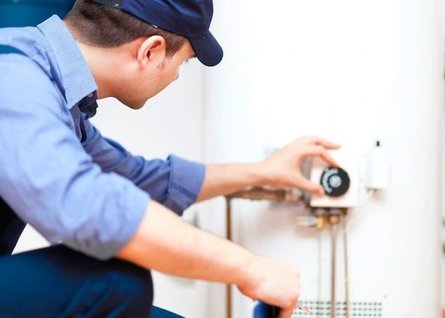 Un técnico de servicio con uniforme azul ajustando el dial de temperatura de un calentador de agua blanco.
