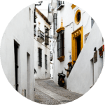 Callejón estrecho y blanco con camino empedrado, puerta amarilla y pequeños balcones en un marco circular.