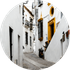 Callejón estrecho y blanco con camino empedrado, puerta amarilla y pequeños balcones en un marco circular.