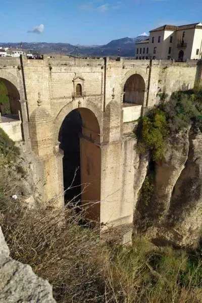 Puente de piedra que cruza un profundo desfiladero, con un edificio histórico en un extremo y montañas en la distancia.