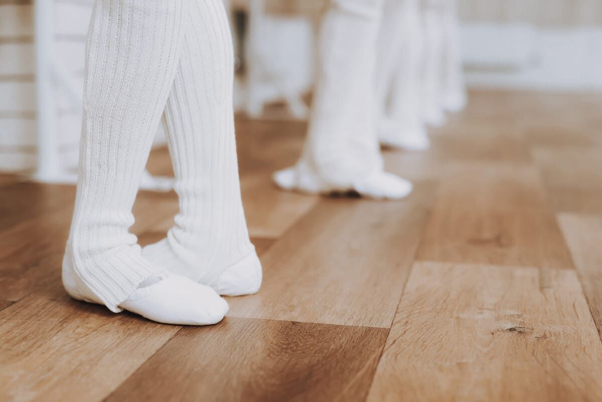 Pies con calcetines blancos sobre el suelo de madera de un estudio de danza, con otros bailarines difuminados al fondo.