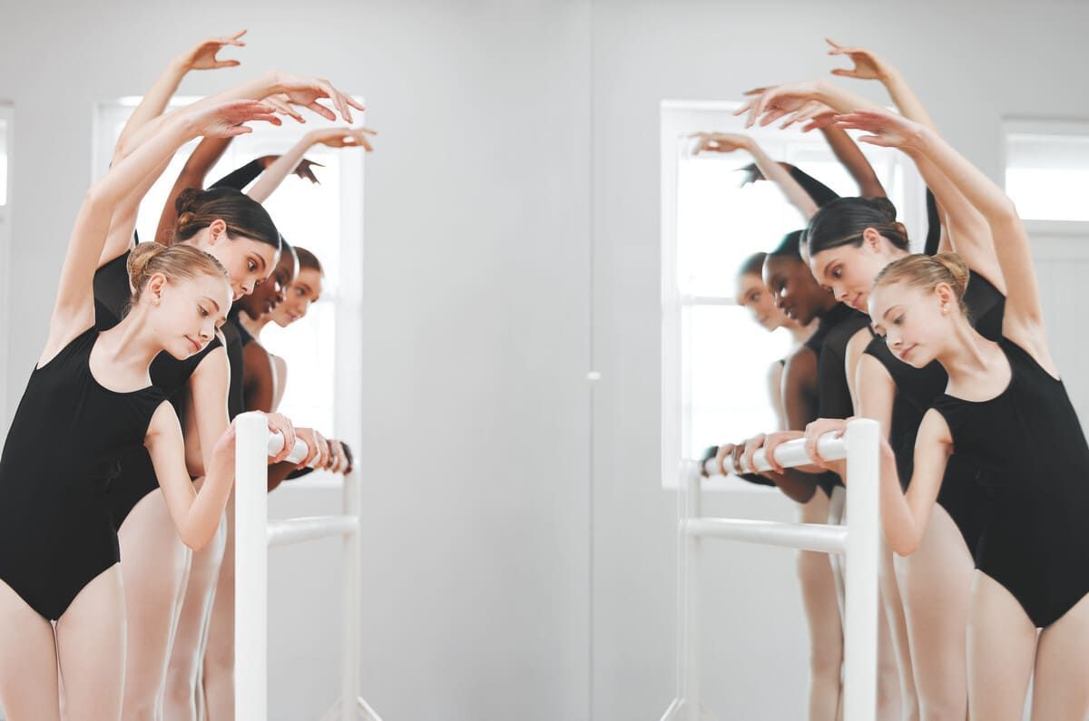 Dos bailarinas con leotardos negros practican en una barra de ballet con los brazos arqueados por encima de la cabeza en un estudio luminoso.