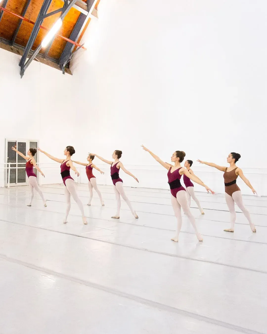 Bailarinas de ballet con leotardos color granate practicando en un estudio luminoso, con los brazos extendidos al unísono.