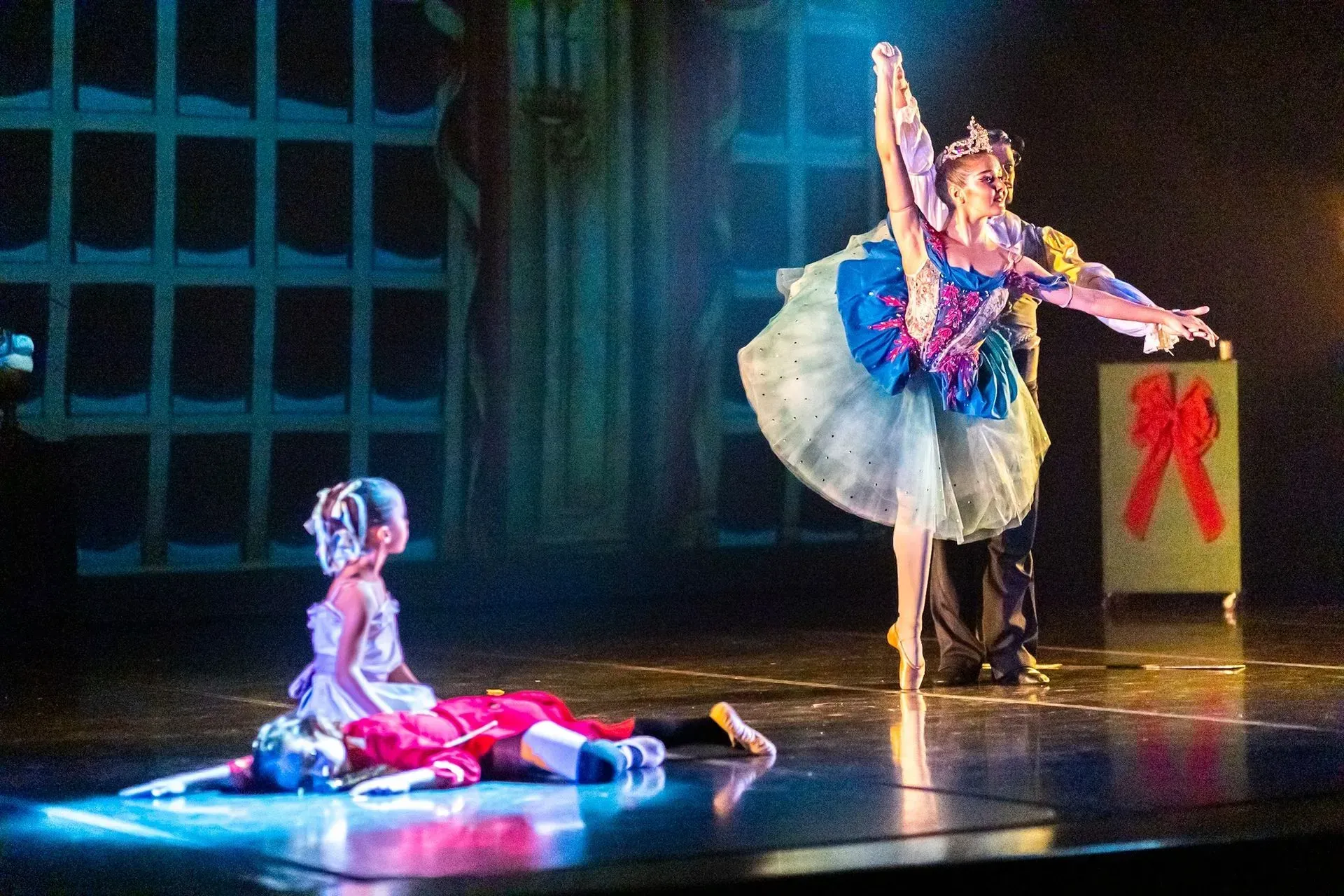 Dos bailarinas de ballet en el escenario, una arrodillada con un vestido rosa y la otra con un tutú azul posando junto a una caja de regalo.