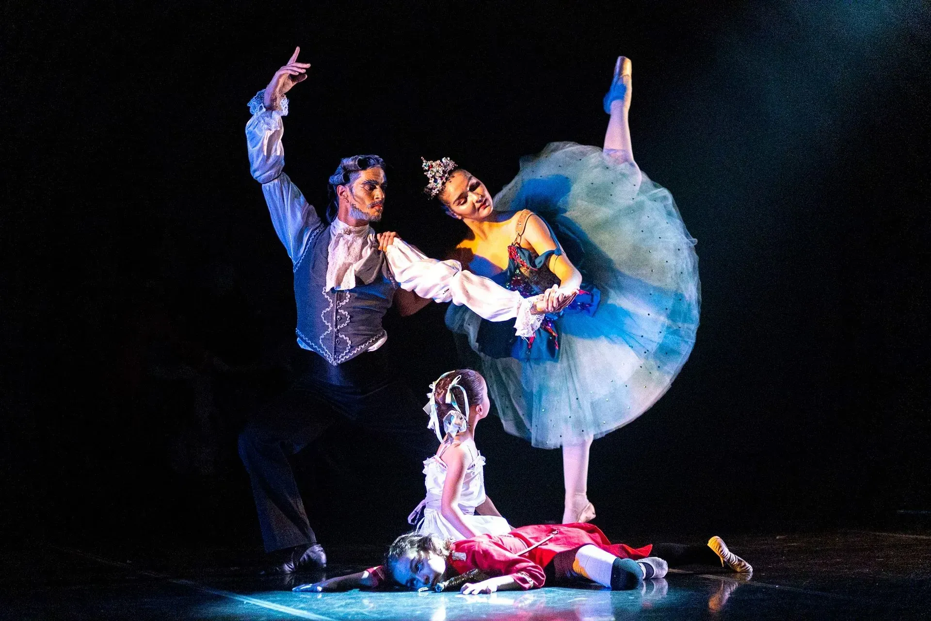 Tres bailarinas de ballet en un escenario oscuro, una con un tutú azul en posición intermedia, una sentada con un vestido rojo, una saltando