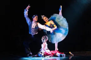Tres bailarinas de ballet en un escenario oscuro, una con un tutú azul en puntas y dos sentadas debajo.