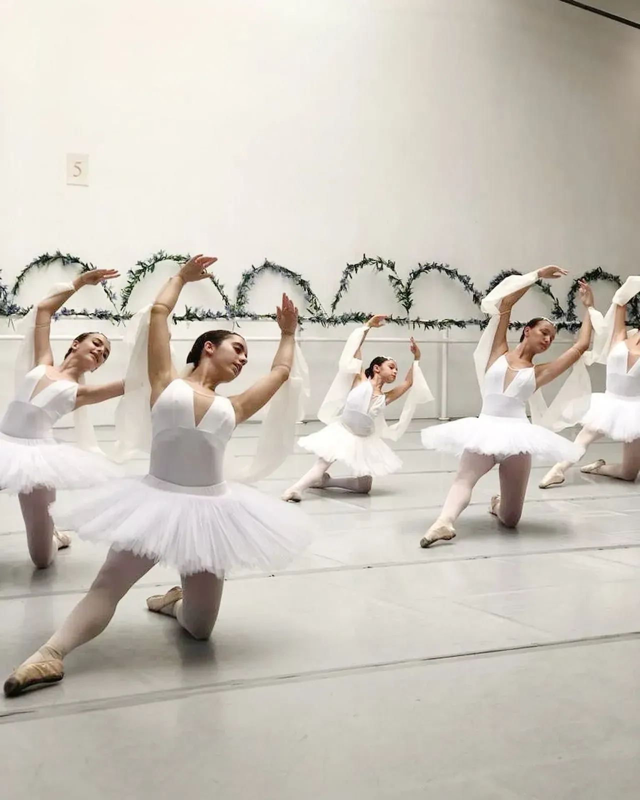 Bailarinas de ballet con tutús blancos realizando una pose sincronizada en un estudio con guirnaldas de corazones en la pared.