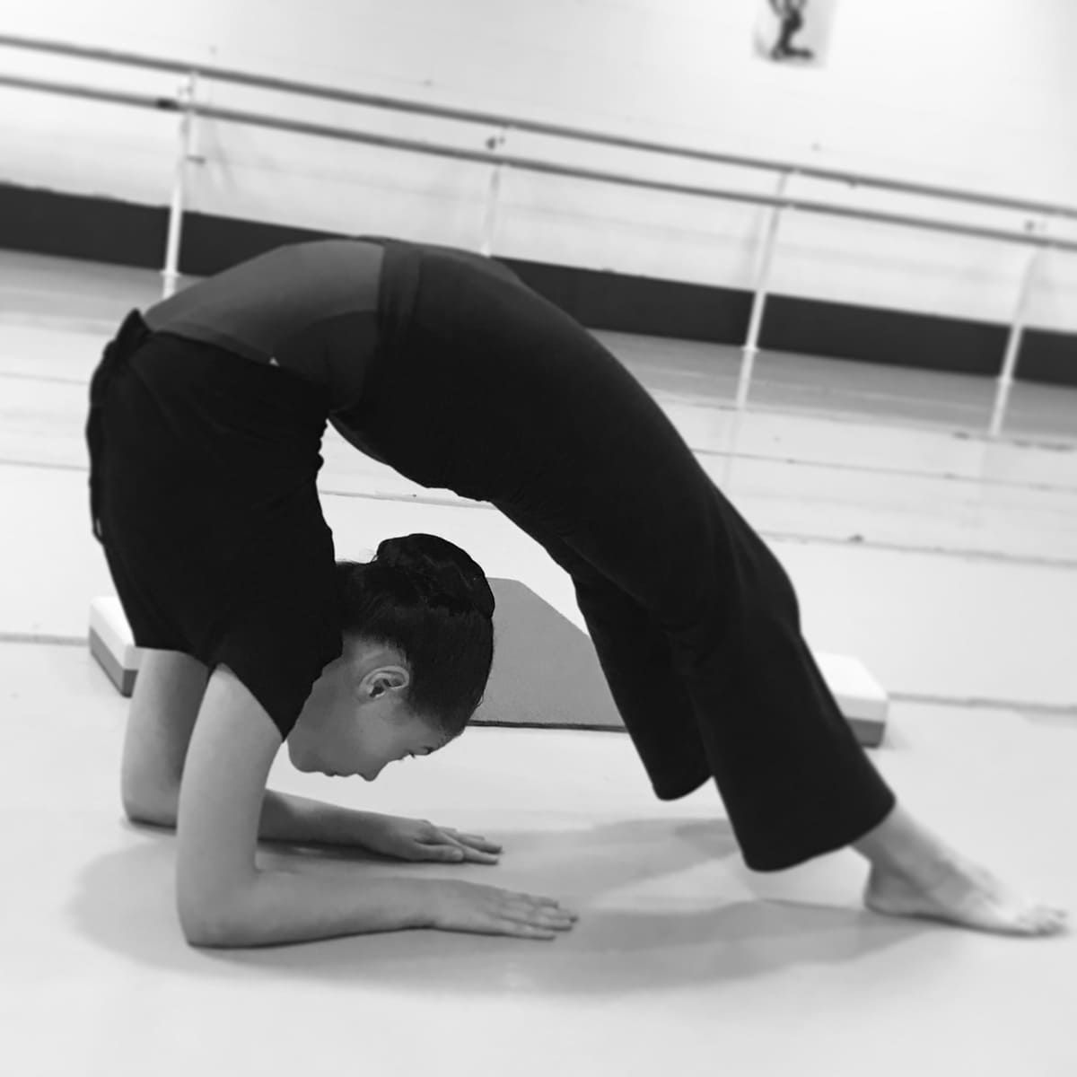 Una persona vestida de negro realiza un estiramiento profundo de espalda en un estudio de ballet con una barra.