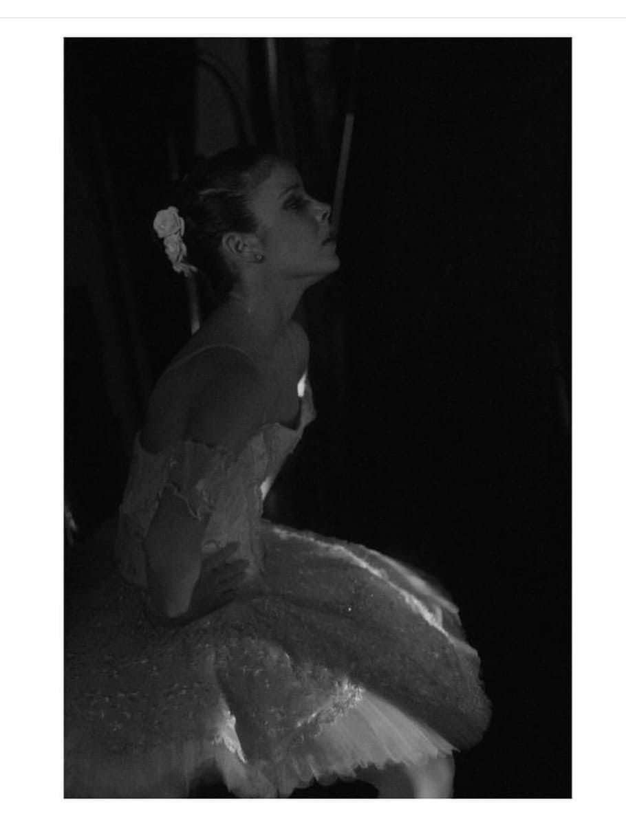 Fotografía en blanco y negro de una bailarina de perfil, sentada con un tutú, sobre un fondo oscuro.