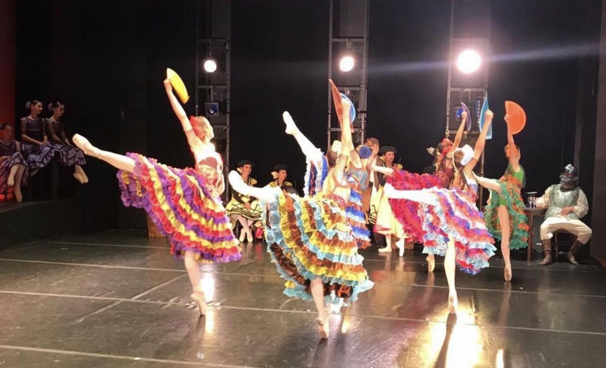 Bailarines con coloridos trajes interpretan un ballet sincronizado en el escenario bajo focos.