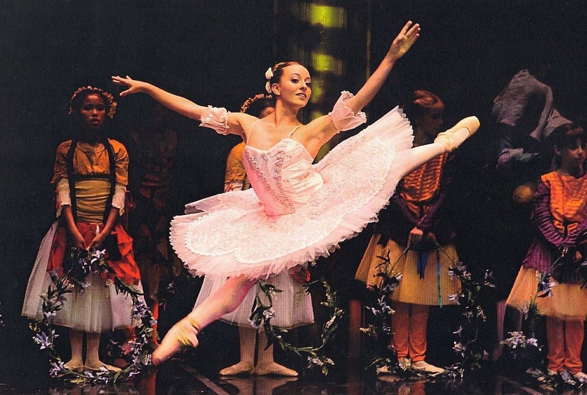 Una bailarina con tutú rosa realiza un grand jeté en el escenario, con otras bailarinas de pie detrás de ella.