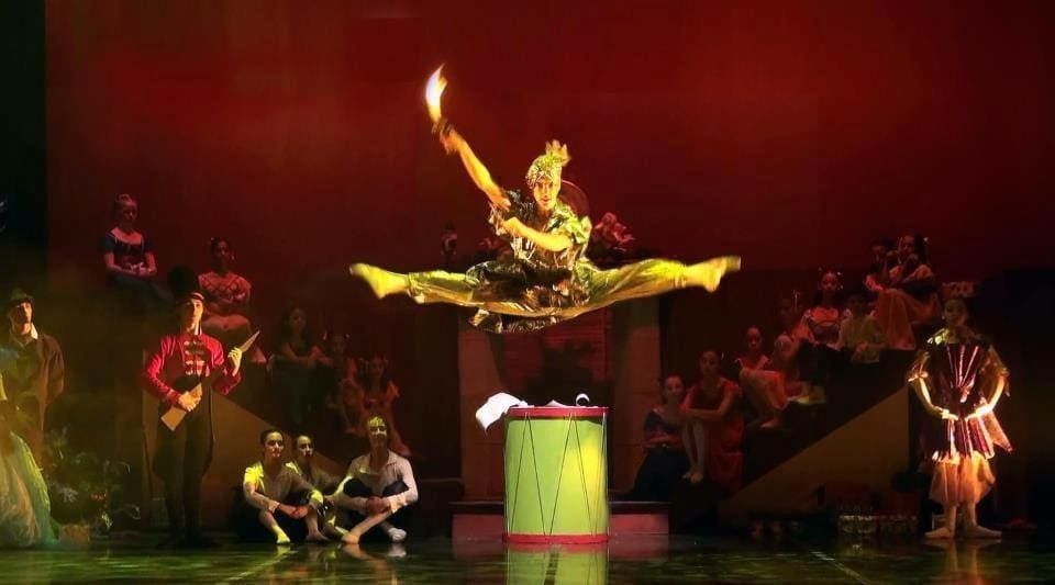 Un artista escénico con traje dorado se equilibra sobre un pedestal con el brazo levantado, con bailarines iluminados con luz roja oscura detrás.