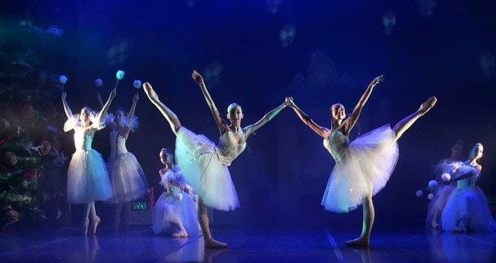 Bailarinas de ballet con tutús blancos actúan en un escenario iluminado con luz azul, con los brazos en alto.