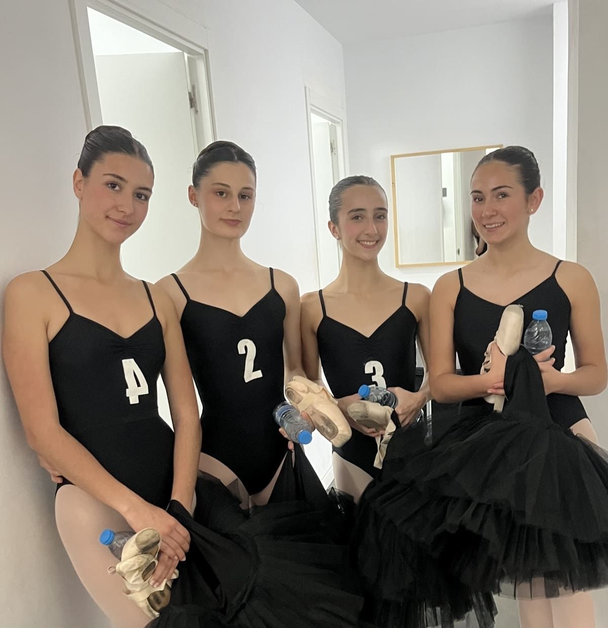 Cuatro bailarinas con trajes negros sosteniendo zapatillas de punta en un luminoso camerino.