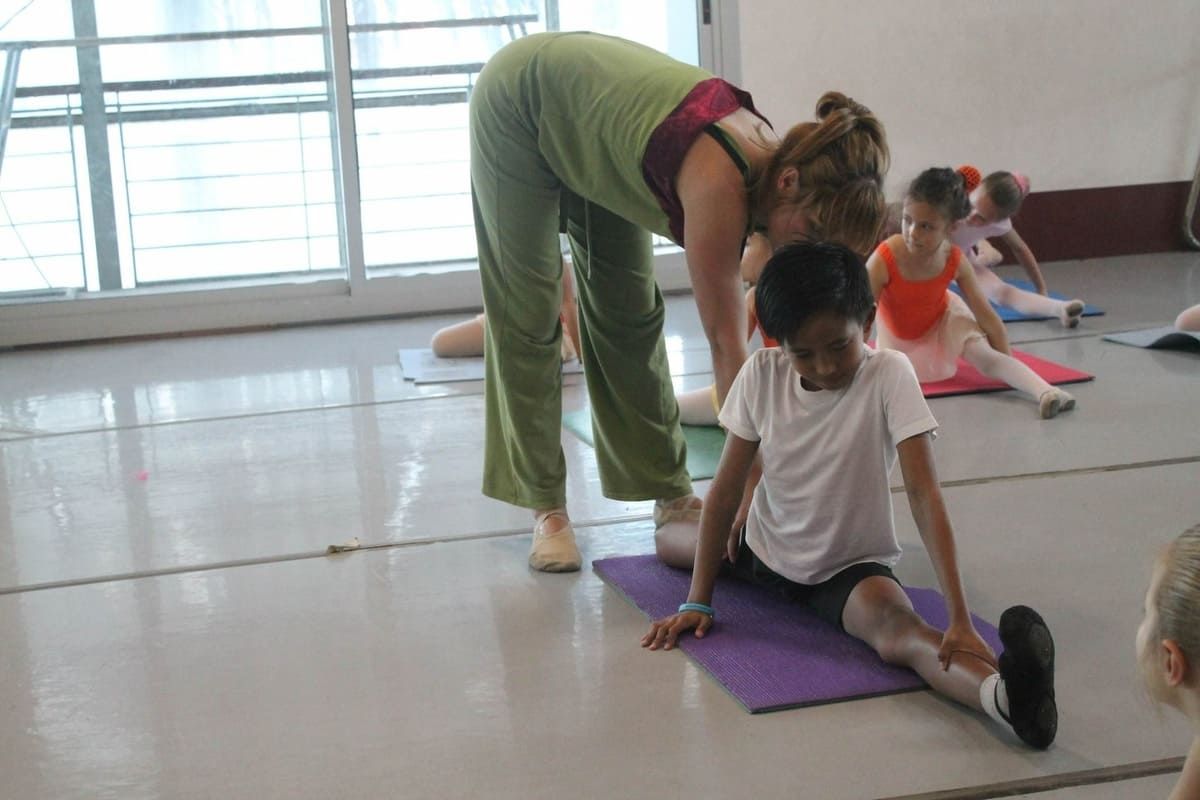 La instructora ayuda a un niño a estirarse sobre una colchoneta morada en un luminoso estudio de danza, mientras otros niños se estiran cerca.