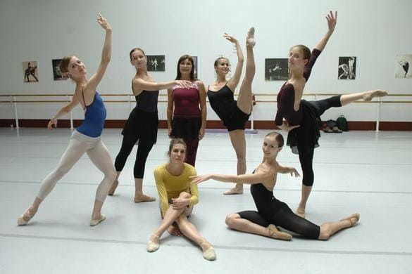 Siete bailarinas de ballet posando en un estudio, vestidas con ropa de ensayo de color negro, blanco, azul y rojo.