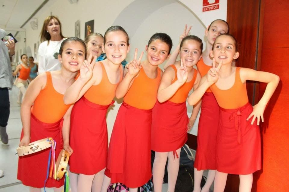 Seis niñas con blusas naranjas y faldas rojas posan sonriendo en un pasillo, con un adulto al fondo.