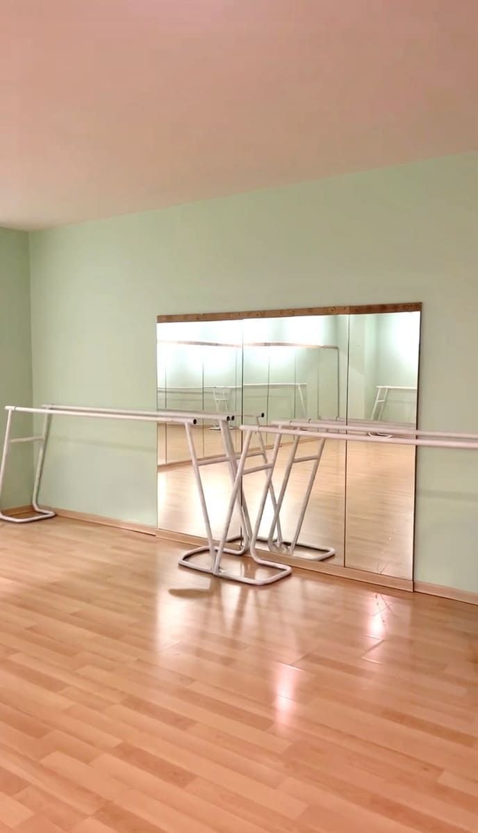 Estudio de danza vacío con suelo de madera noble, paredes de color verde pálido y barras de ballet reflejadas en una de las paredes.