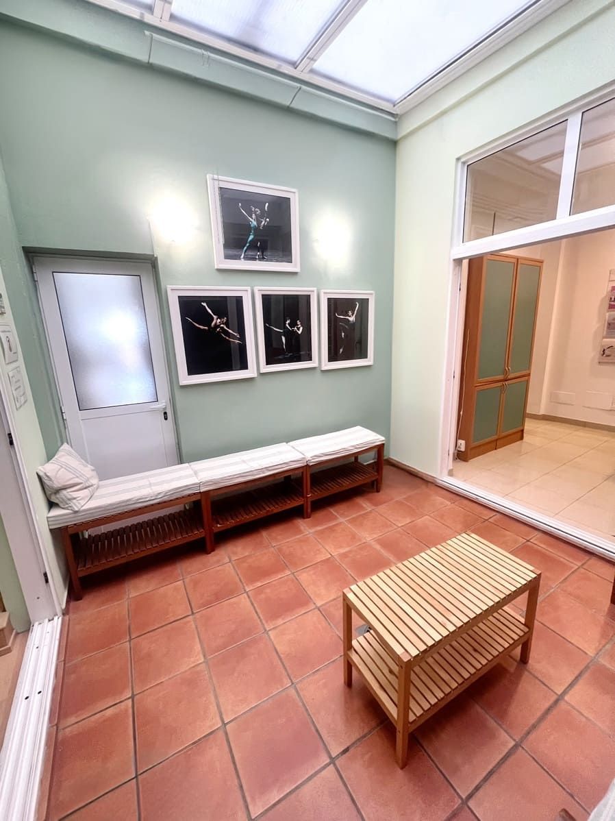Pequeña sala de espera con suelo de baldosas, banco, mesa baja, decoración en la pared y puerta abierta bajo un tragaluz.