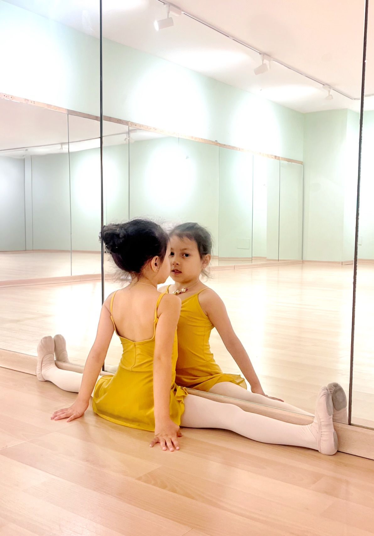 Niña con traje de ballet amarillo sentada en un estudio de danza, frente a una pared con espejo.