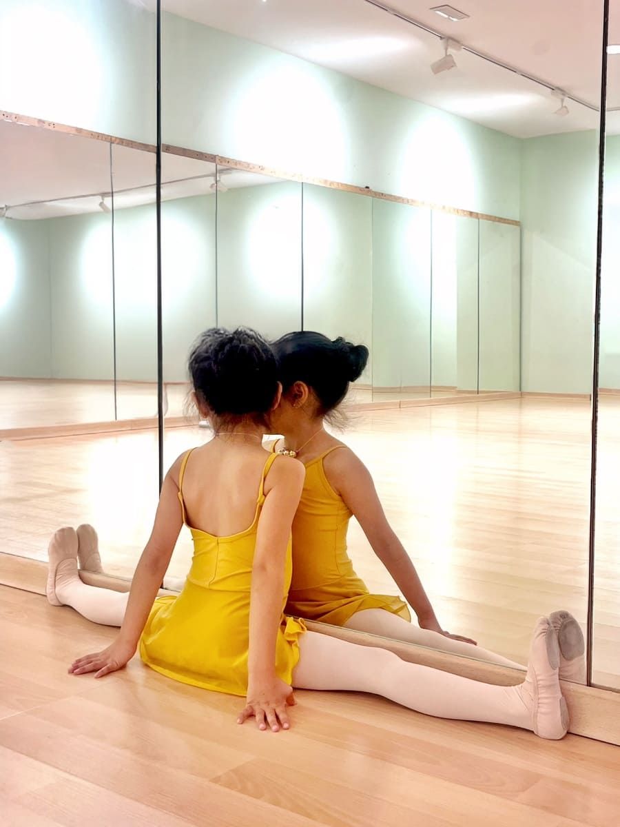 Una persona vestida con un vestido amarillo está sentada en un estudio de danza, estirándose contra una pared con espejos.