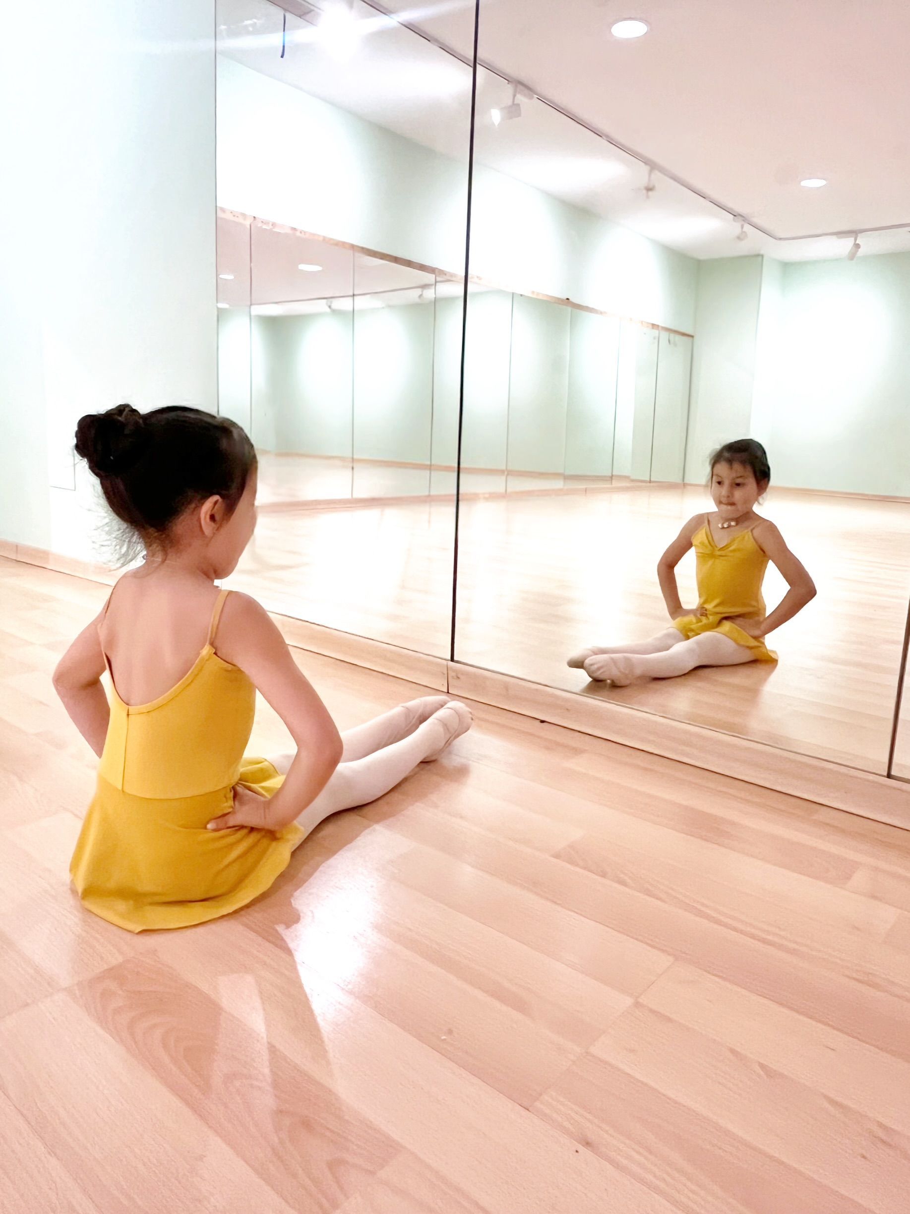 Niña con leotardo amarillo sentada en el suelo de un estudio, frente a una pared con espejo.
