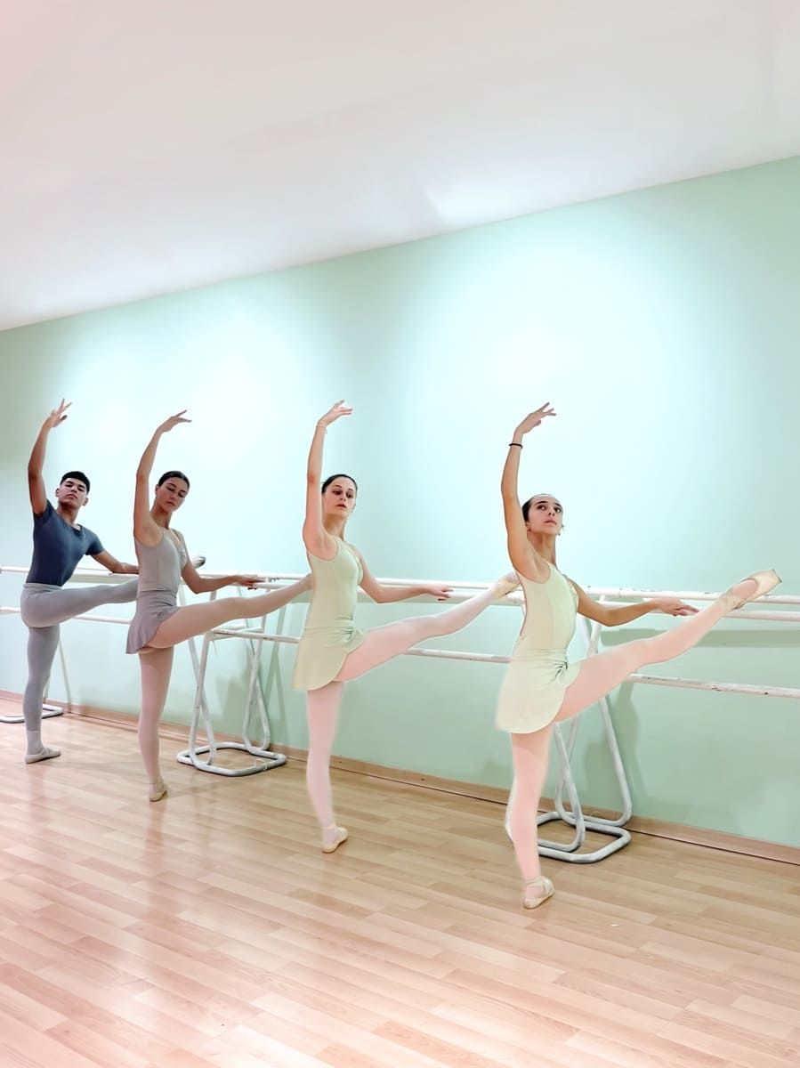 Cuatro bailarinas con leotardos mantienen posturas de arabesco en un luminoso estudio de ballet con suelo de madera.