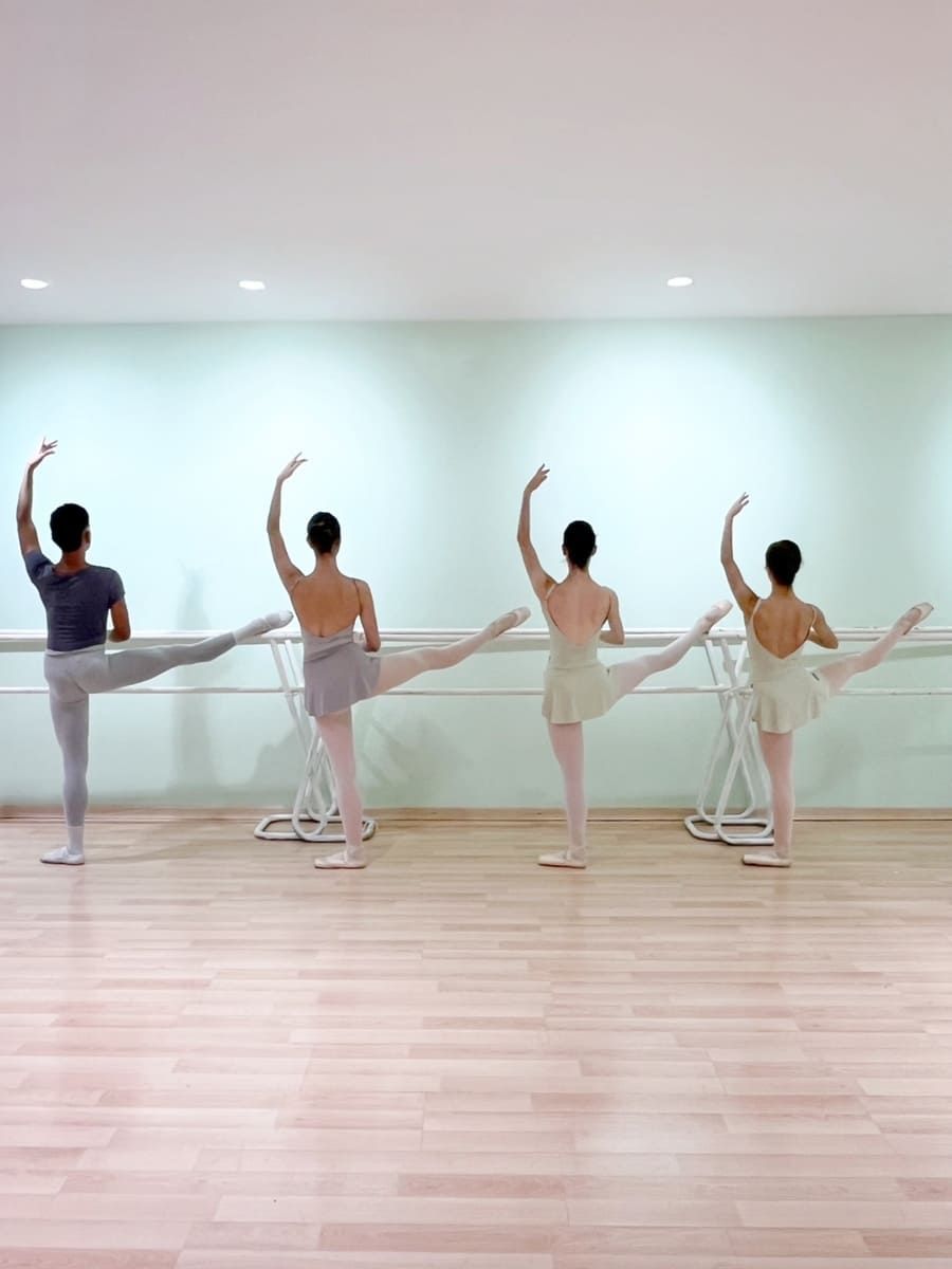 Cuatro bailarinas en un estudio mantienen posturas de equilibrio de ballet en una barra, con las piernas extendidas y los brazos levantados.