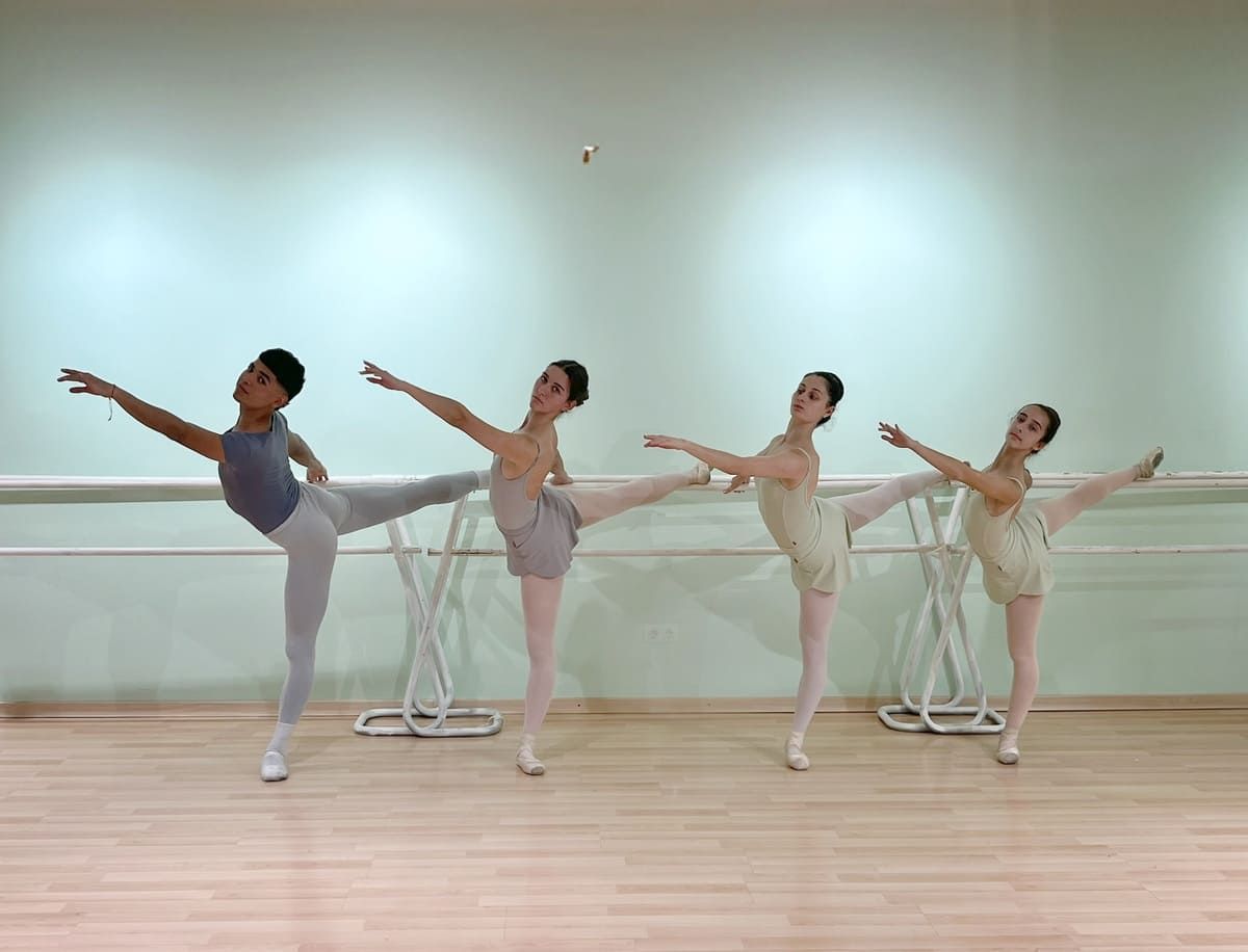 Cuatro bailarinas de ballet con leotardos de colores pastel practican arabescos en un estudio con espejos y suelo de madera.