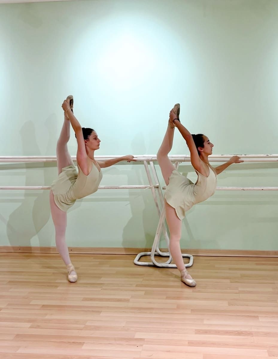 Dos bailarinas de ballet con tutús beige posan en un estudio, cada una manteniendo una extensión de pierna alta en la barra.