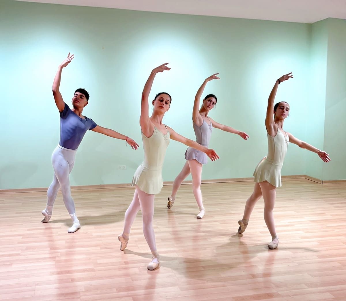 Cuatro bailarinas de ballet con trajes en tonos pastel posan en un estudio luminoso con suelo de madera.