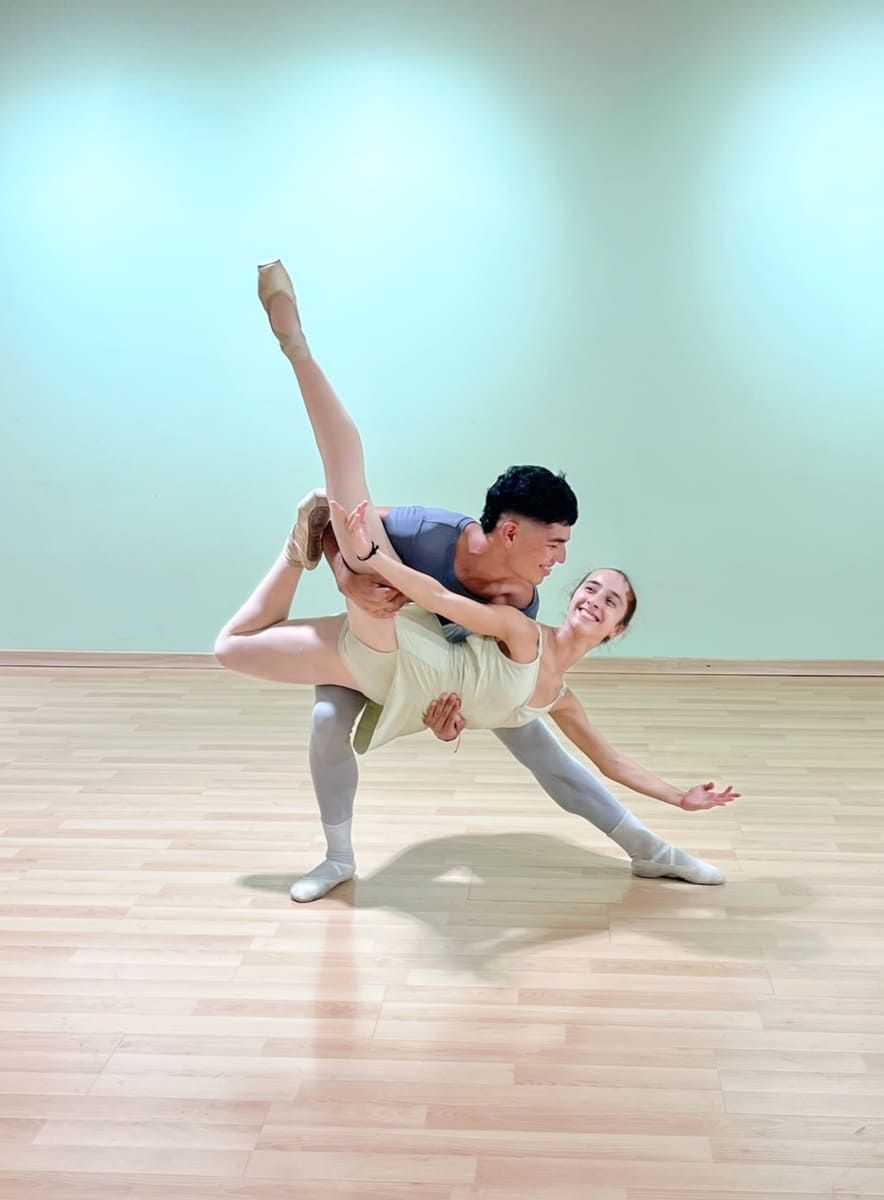 Dos bailarinas de ballet en una pose de elevación dramática sobre un suelo de madera clara en un estudio con una pared azul de fondo.