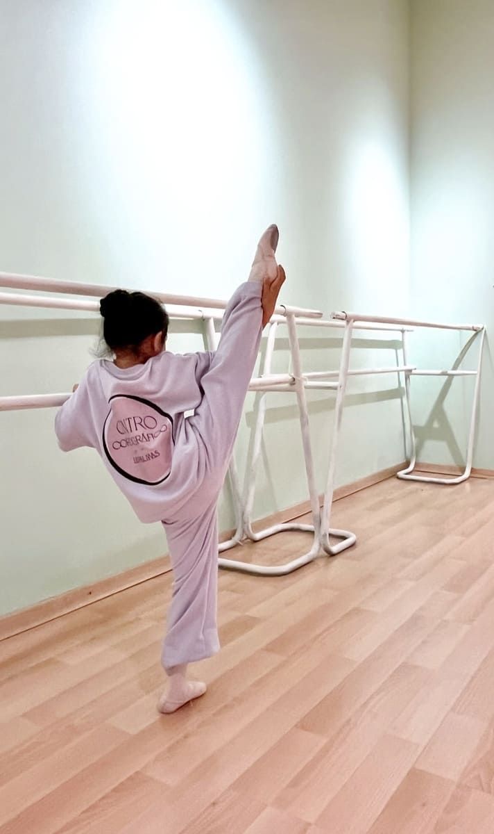 Una persona con un chándal rosa pálido realiza estiramientos en una posición de pierna alta en un estudio de ballet con barra y suelo de madera.