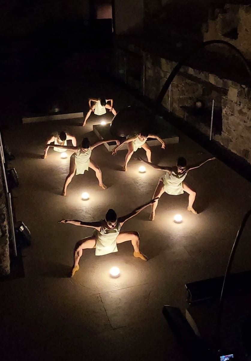 Personas haciendo una postura de yoga o baile en un suelo tenuemente iluminado alrededor de luces que parecen velas.