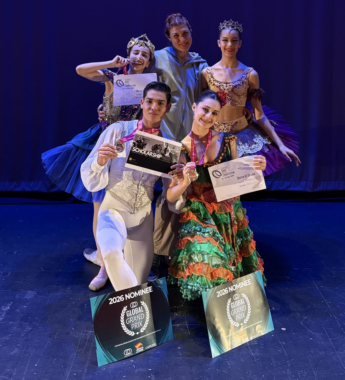 Cinco bailarines posan en un escenario con certificados y bolsas de premios bajo luces azul-violeta.