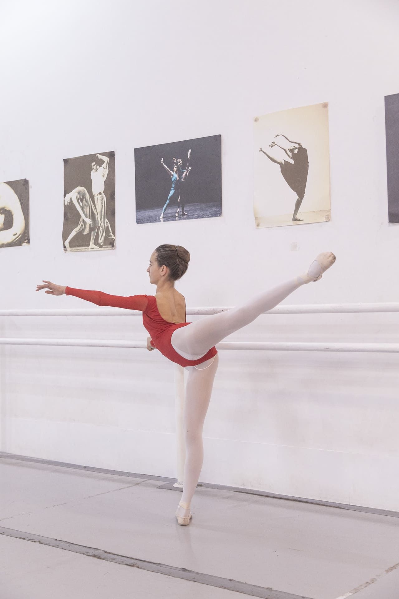 Una bailarina de ballet con leotardo rojo realiza una extensión de pierna alta en una galería de un estudio.