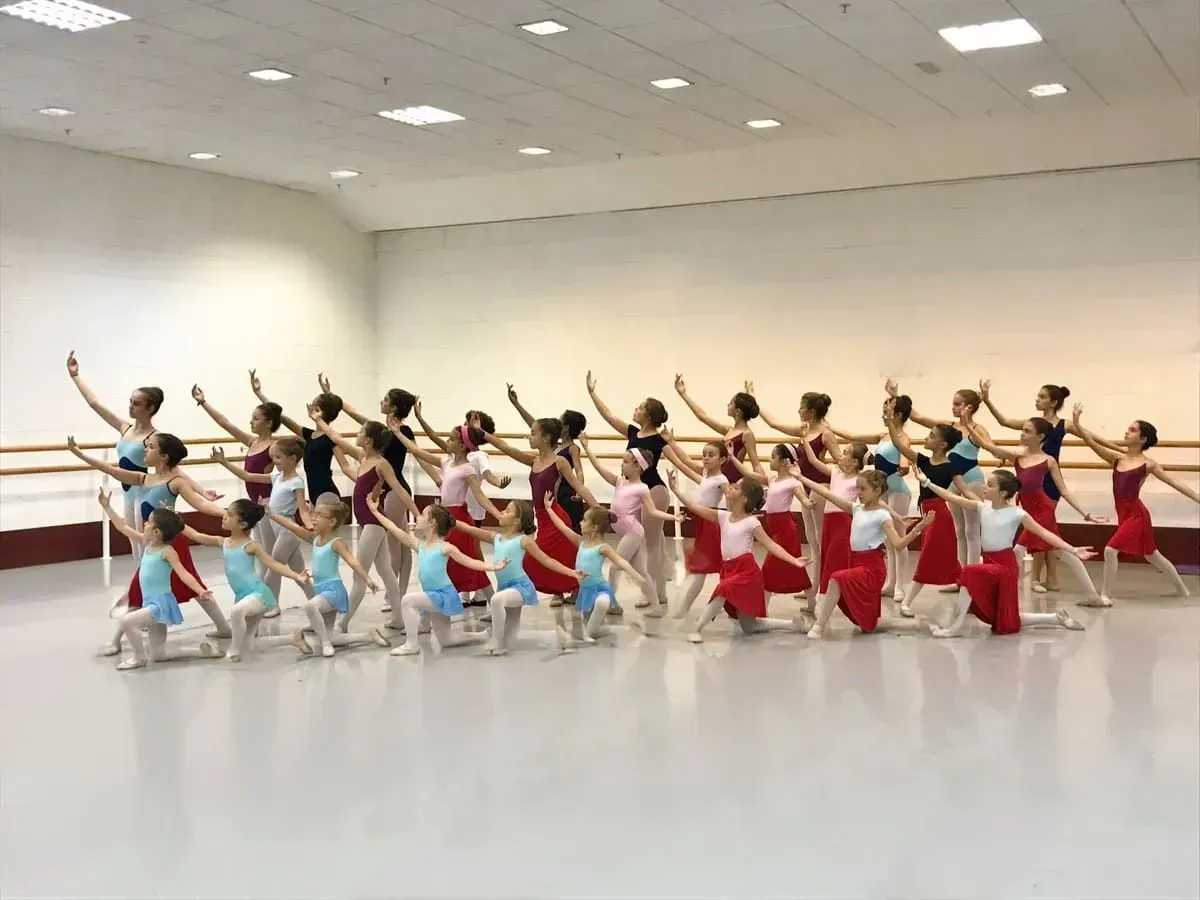 Bailarines de ballet con coloridos trajes posan en un estudio luminoso con los brazos levantados al unísono.