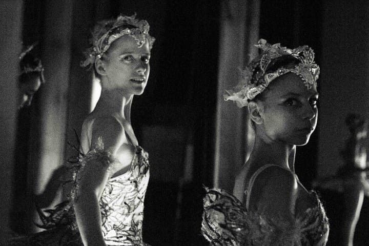 Dos mujeres con tocados florales y vestidos brillantes permanecen de pie en una habitación con poca luz, mirando por encima del hombro.