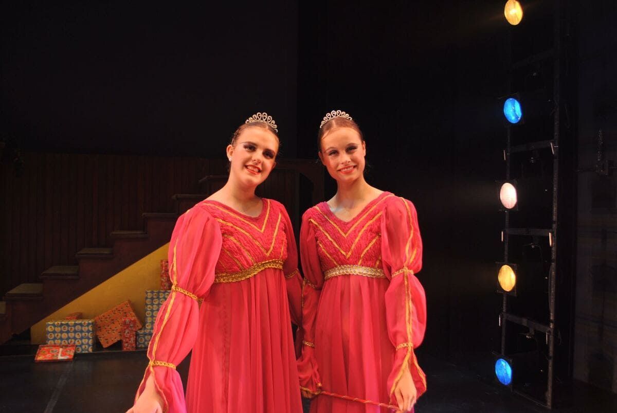 Dos artistas con vestidos de color rosa brillante están de pie en un escenario tenuemente iluminado con luces de colores a su lado.